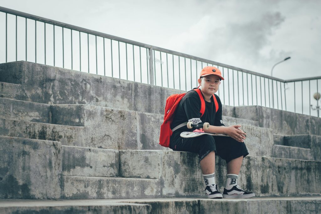 A young boy sitting on concrete steps with a skateboard, wearing casual attire and a red backpack.
